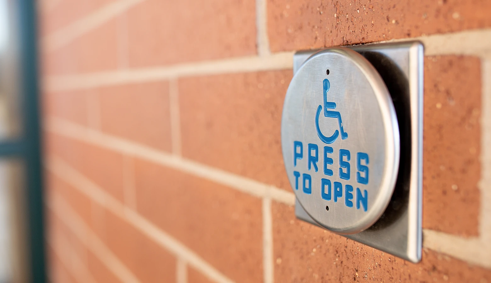 A sign on a brick wall that says "Press to Open", with the international accessibility symbol.